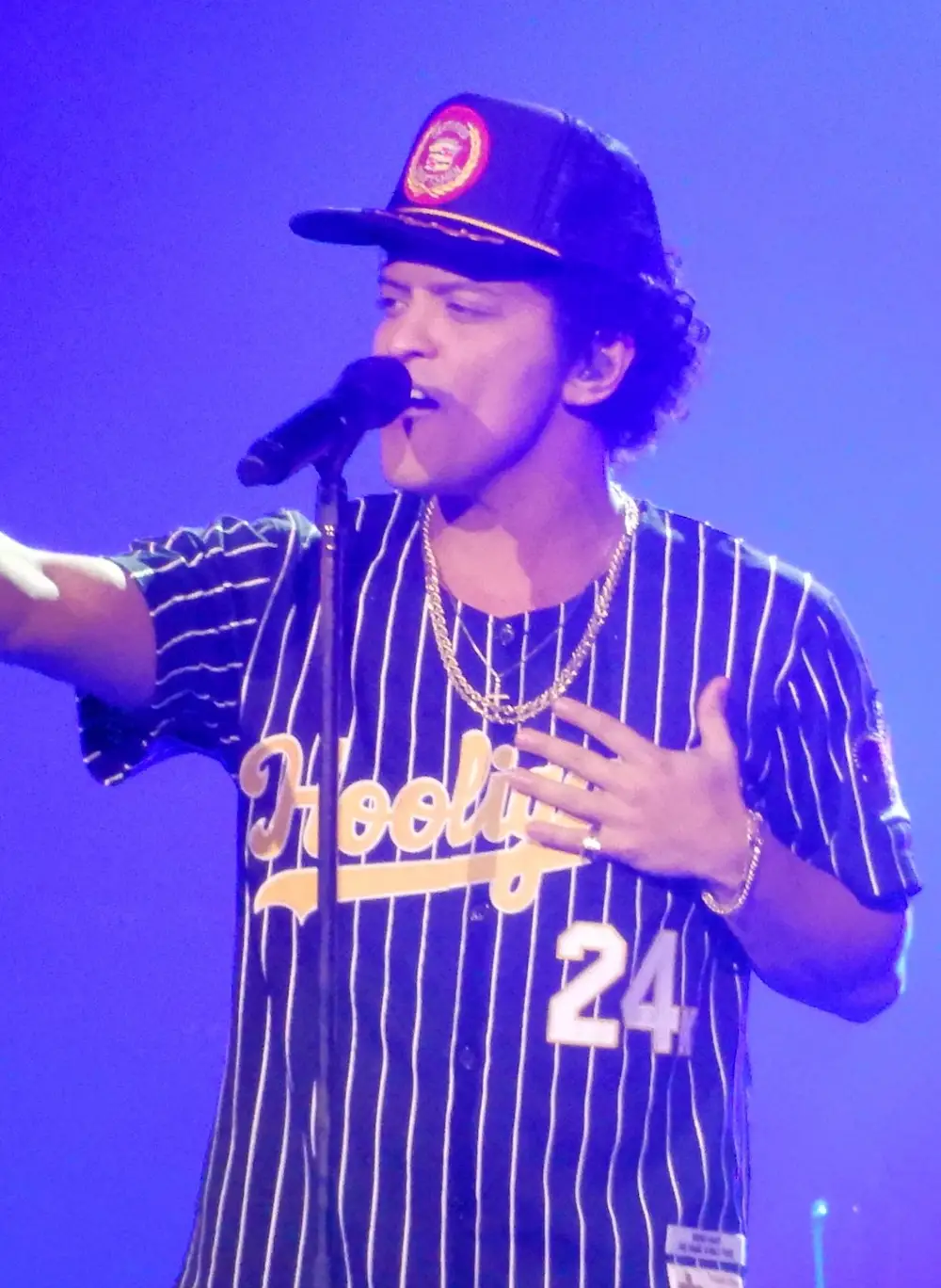 Bruno Mars cantando ao vivo ao microfone na turnê 24K Magic, com boné e camisa listrada