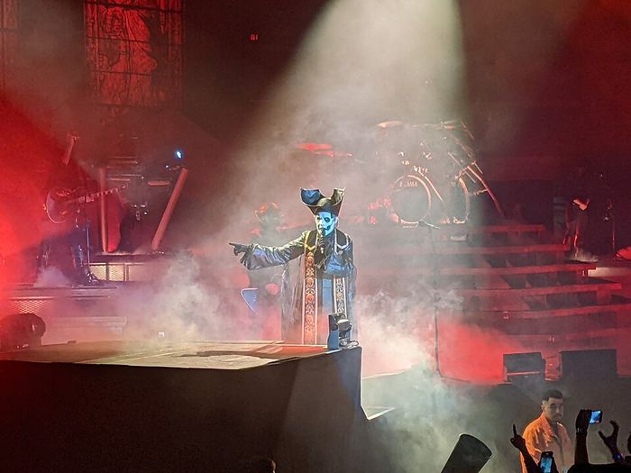 Silhueta de vocalista com as mãos erguidas diante de luz forte no palco, em pose quase sacerdotal